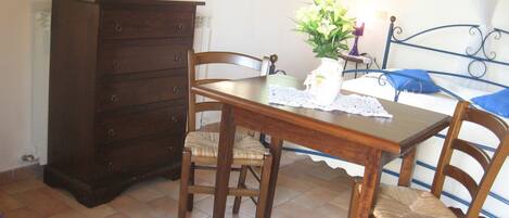 Una habitación con una mesa de madera, dos sillas, un aparador, una cama con cabecera azul y una ventana con cortinas.