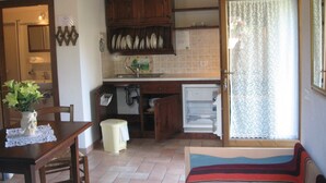 Une cuisine de taille réduite, équipée d’un évier, de placards et d’un réfrigérateur. On y trouve une table à manger avec des chaises et un petit tabouret. Une fenêtre avec des rideaux est visible, et une porte donne accès à une autre pièce.