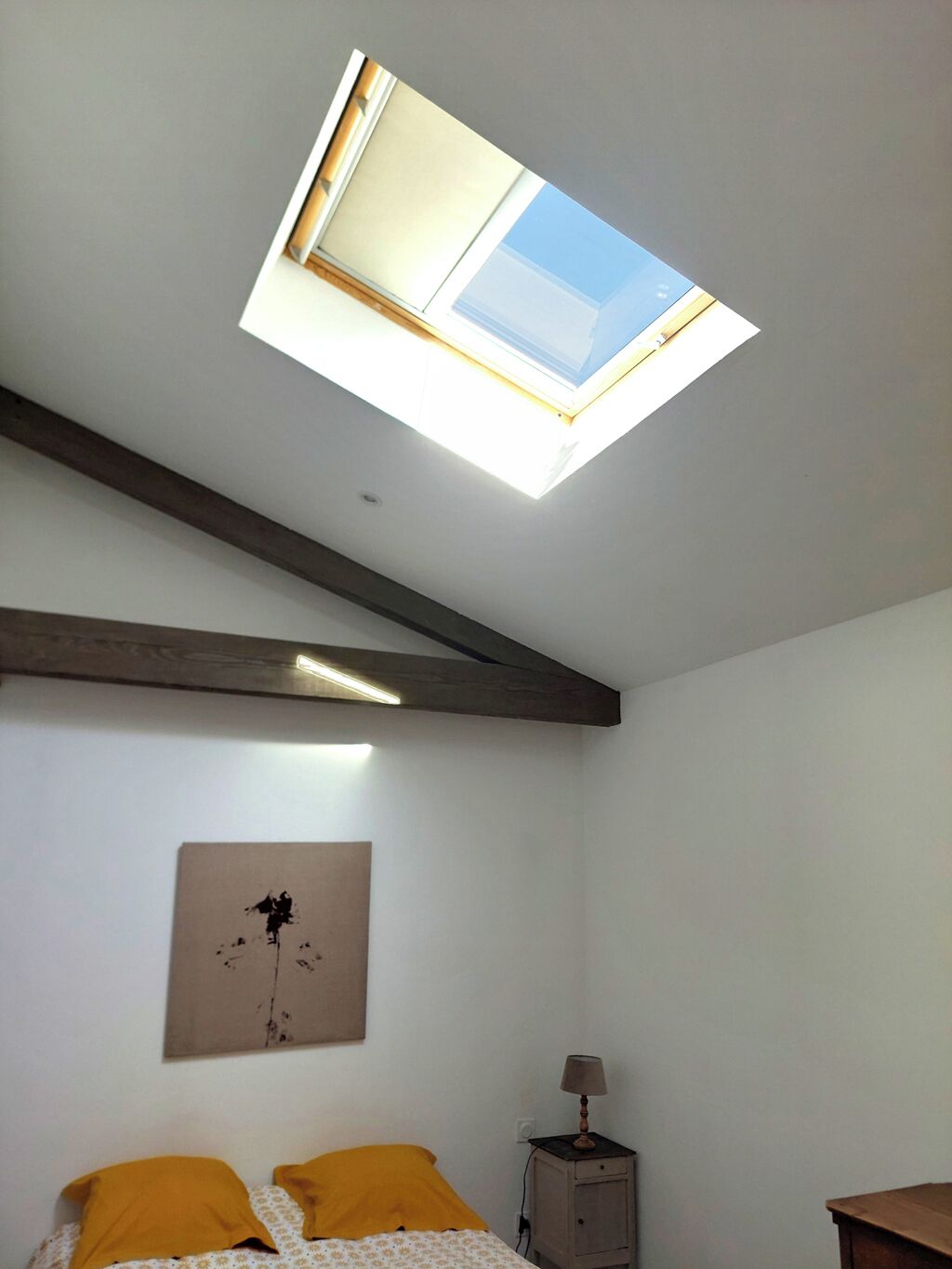 A bedroom with a skylight, a bed with yellow pillows, a nightstand, and a wall-mounted lamp.