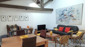 A living room with a wooden dining table, chairs, a sofa with cushions, a bookshelf, a flat-screen TV, and a large abstract painting on the wall. - LA Magdeleine (Cabannes)