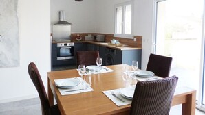 A dining area with a wooden table set for four, a kitchen in the background, and a large window providing natural light. - LA Magdeleine (Cabannes)