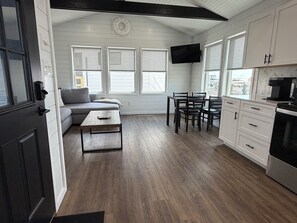 Cocina moderna con muebles blancos, zona de comedor con mesa y sillas, televisor empotrado y un amplio ventanal.