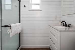 Un baño con paredes de madera blanca, cabina de ducha de vidrio, lavamanos blanco con grifo negro y un pequeño ventanal sobre el lavamanos.