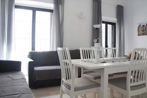Un coin repas avec une table et des chaises blanches, un canapé noir et une fenêtre avec des rideaux.