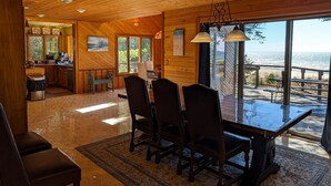 A spacious dining area with a large table and chairs, a kitchen with wooden cabinets, and a view of the ocean through large windows.
