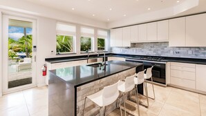 Una cucina moderna con una grande isola, mobili bianchi e vista su un giardino tropicale.