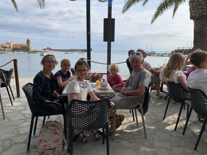 A group of people seated at an outdoor table with a view of the sea and a historic building in the background.
