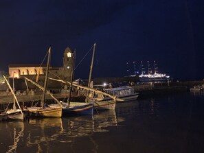 A harbor at night with boats docked, a building with a tower, and a large ship in the background.