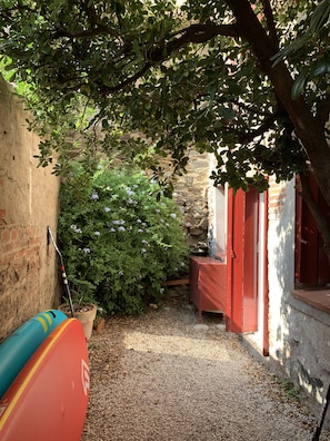 A narrow alleyway with a gravel path, a red bench, and a surfboard leaning against a wall.