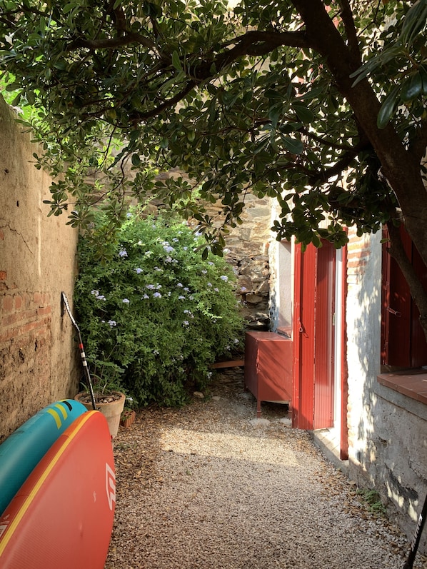 A narrow alleyway with a gravel path, a red bench, and a surfboard leaning against a wall.