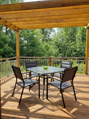 A covered outdoor deck with a table and chairs, surrounded by a wooden railing and lush greenery in the background. - Condo on Golf near Bike Trail w/ 2 Bdrm 2 Bath (Mont-Tremblant)