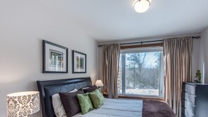Une chambre avec un grand lit, une table de chevet avec une lampe, une commode et une fenêtre avec des rideaux.