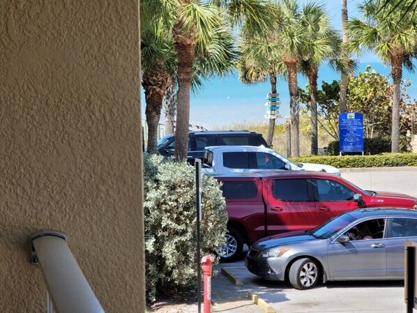 Un espace de stationnement avec un SUV rouge et une voiture argentée, des palmiers et un panneau d’information bleu.