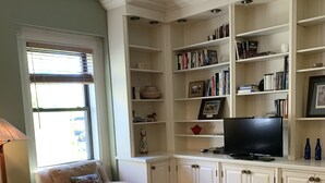 Sala de estar con un sofá de cuero marrón, un sillón color beige y un televisor sobre un mueble bajo. Se observan estanterías con libros y objetos decorativos.