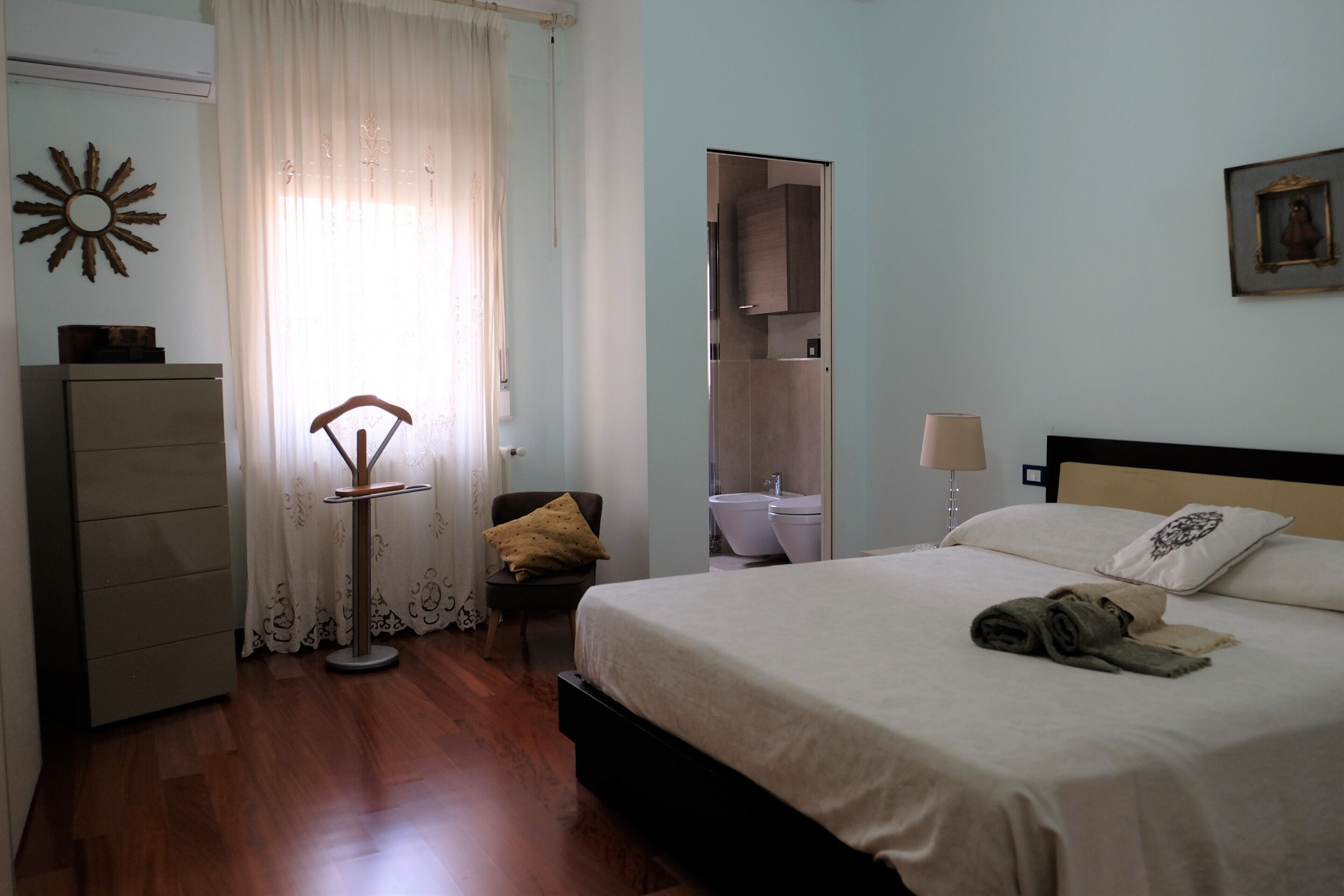 Un dormitorio con cama, una silla, un escritorio y un baño visible a través de una puerta abierta.