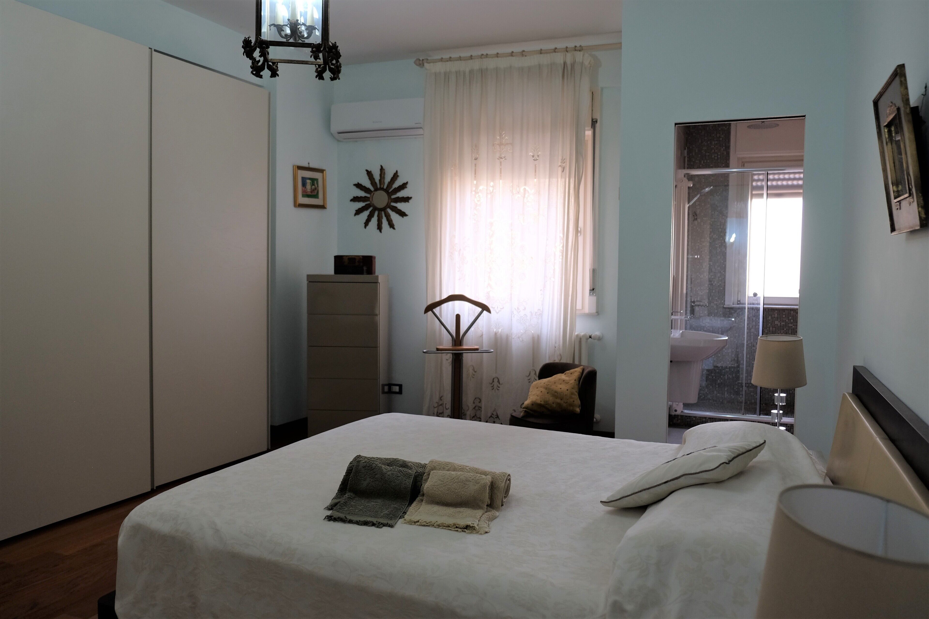 Un dormitorio con cama, armario, ventana con cortinas, mesita de noche y lámpara.