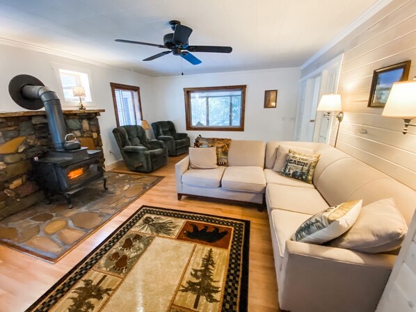 A cozy living room with a stone fireplace, a sofa, two armchairs, a ceiling fan, and a rug with a forest design. - Cozy Bear Den - private house with fenced yard! (Seeley Lake)