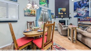 A dining area with a round table, chairs, and a chandelier. There is a fireplace and artwork on the walls.