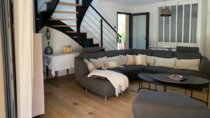 Un salon moderne avec un canapé d’angle, une petite table ronde, un escalier doté d’une rampe noire et un luminaire suspendu.