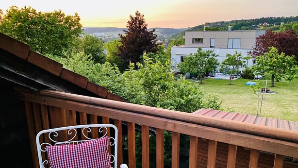 A balcony with a wooden railing, a white metal chair with a checkered cushion, and a view of a green landscape with trees and a building in the background.