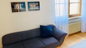 A living room with a grey sofa, two framed pictures on the wall, a window with sheer curtains, and a radiator.
