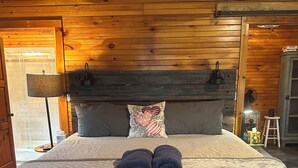 Camera da letto con rivestimento in legno, un letto, cuscini, una lampada da comodino e un piccolo tavolo con lampada.