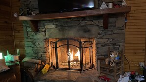 Ein Kamin aus Stein mit einem Fernseher darüber, eine Holzbank und ein Stofftier auf einem Stuhl.