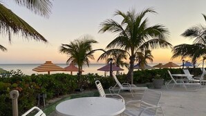 Een terras aan het strand met witte metalen stoelen en tafels, palmbomen en parasols.
