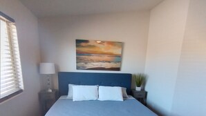 A neatly made bed with a blue headboard, a wall-mounted painting, a bedside lamp, and a small potted plant. - Close to shopping and freeway access.  (Yuma)
