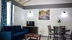 Un salon avec un canapé bleu, une table basse en bois, un téléviseur posé sur un meuble en bois, une table à manger avec des chaises et un tableau accroché au mur.