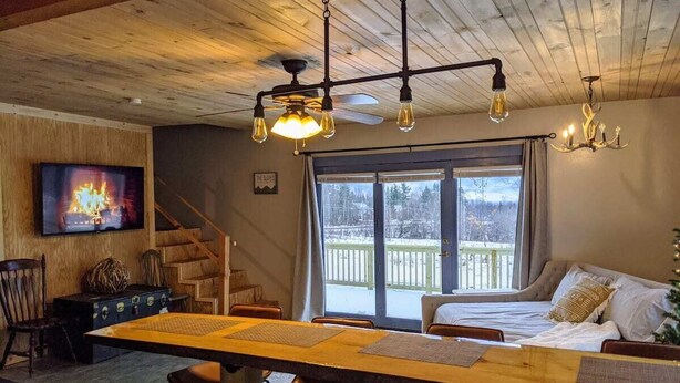 A cozy cabin interior with a wooden dining table, a sofa, a TV, and a staircase leading to a balcony.