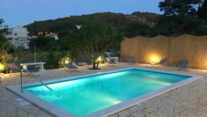 Une piscine rectangulaire bordée d’une terrasse en pierre, entourée de chaises longues et d’une clôture en bois.