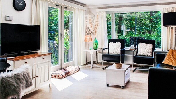 Un salón moderno con un televisor de pantalla plana, dos sillones negros con cojines blancos, una mesa de centro blanca y vistas a zonas verdes a través de amplios ventanales.
