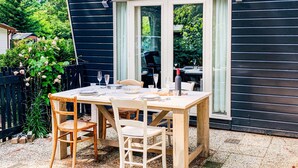 Mesa de madera dispuesta para dos personas con sillas, ubicada en un patio de grava junto a un edificio de madera oscura con ventanas amplias.