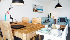 A modern living room with a wooden dining table, a laptop, a plush white chair, a blue sofa, and a painting of a lighthouse. - Beach front apartment in Büsum with sauna (Büsum)