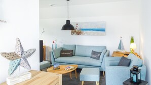 A modern living room with a light blue sofa, matching armchair, and ottoman. There's a wooden coffee table with books and a decorative starfish. A painting of a lighthouse is on the wall, and a small ship model is on a side table. - Beach front apartment in Büsum with sauna (Büsum)