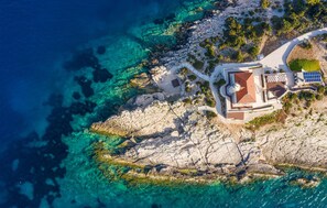 Vue aérienne d’une propriété située sur la côte, avec un bâtiment central, un chemin et un petit jardin.