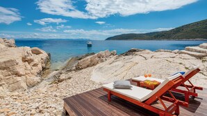 Une terrasse en bois avec un fauteuil de relaxation et une table, d’où l’on peut admirer une mer calme, avec un bateau au loin et un paysage rocheux.