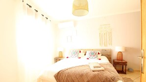 A well-lit bedroom with a large bed, a wooden headboard, bedside tables with lamps, and a hanging macrame decoration.