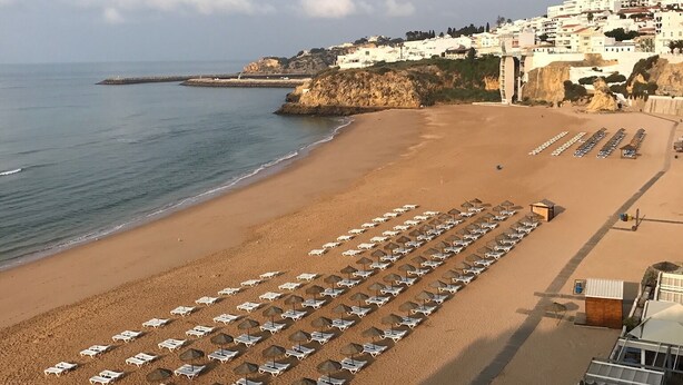 A beach with rows of sun loungers and umbrellas, a rocky coastline, and buildings in the background.
