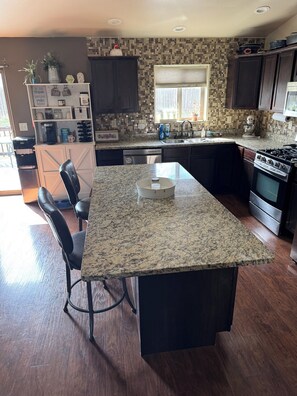 A kitchen with a granite island, dark cabinets, a dishwasher, a stove, and a window with blinds. - Beautiful! (Black Hawk)