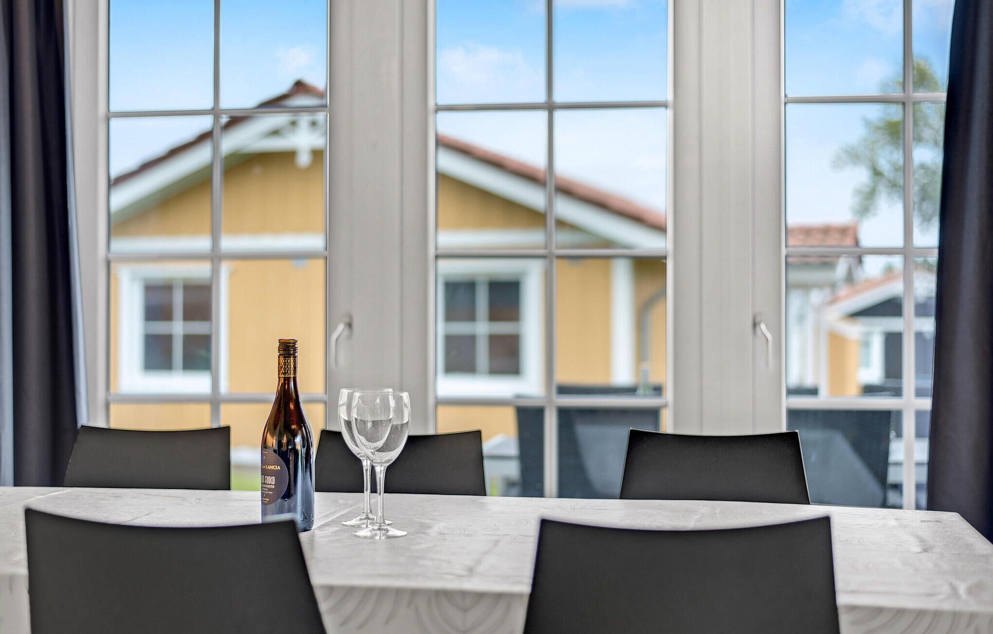 A dining table with a bottle and two glasses, large windows with a view of houses, and a modern interior design.