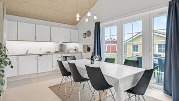 Et moderne køkken med hvide skabe, et spiseområde med et marmorbord og sorte stole, samt store vinduer med gardiner.