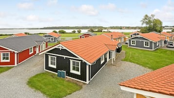 A row of small, single-story cabins with red-tiled roofs, arranged in a row with a grassy area in front and a body of water in the background.