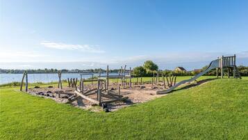 A playground with a slide and swings, set against a backdrop of a lake and grassy area.
