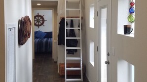 Un pasillo estrecho con piso de madera, una puerta blanca con ventana y una escalera que sube a una zona de cama en un altillo.