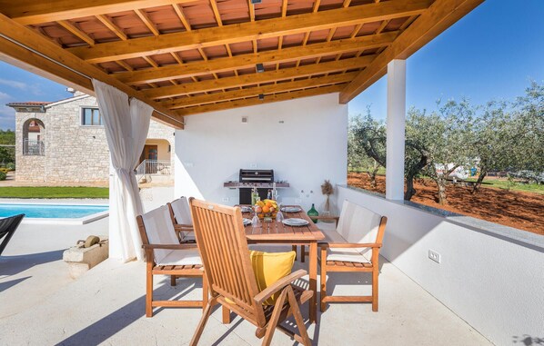 An outdoor patio with a wooden table and chairs, a grill, and a view of an olive grove. - Stunning home in Galizana (Galizana)