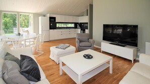 Un salon moderne avec un canapé blanc, une table basse et un téléviseur. On aperçoit une cuisine à l’arrière-plan, avec des meubles blancs et une grande fenêtre.