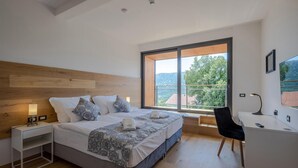 A modern bedroom with a wooden headboard, a large bed with patterned bedding, a bedside table with a lamp, a desk with a chair, and a large window with a view of greenery. - Luxurious home in Grizane with sauna (Grizane)