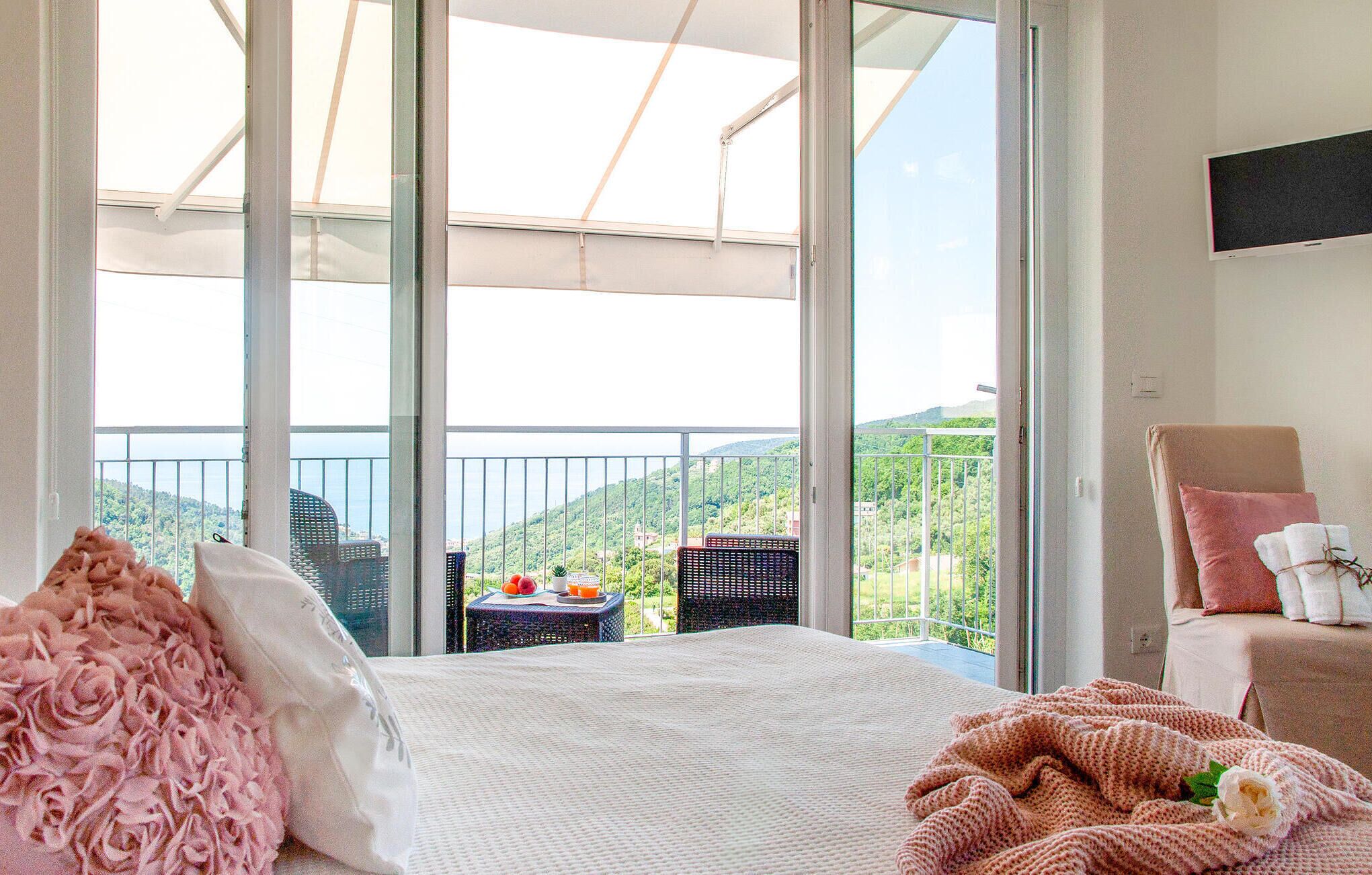 Ein Hotelzimmer mit einem großen Balkon, einem Bett mit rosa-weißer Bettwäsche und einem Sessel mit Decke.